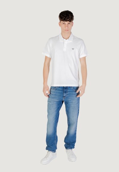 T-shirt polo blanc avec deux boutons, associé à un jean bleu clair. Le modèle porte des baskets blanches, debout sur un fond clair.