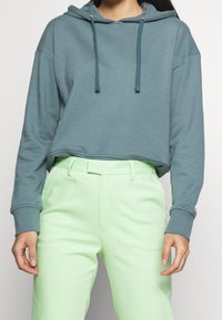 Sudadera corta color verde azulado con capucha y cordones, hecha de tela suave. Combinada con pantalones de talle alto y corte entallado en verde claro.