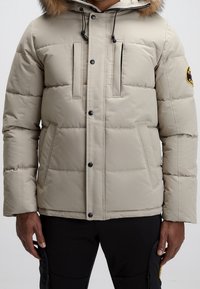 Doudoune beige avec une capuche en fourrure, comportant des coutures horizontales, deux poches avant et des accents en métal noir.