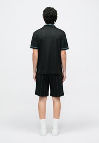 Άνδρας με σγουρά μαλλιά στέκεται με την πλάτη, φοράει μαύρο πόλο με λεπτές ρίγες, μαύρα branded σορτς, λευκές κάλτσες και λευκά αθλητικά παπούτσια σε απλό φόντο.