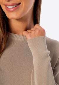 Beżowy sweter dzianinowy z prążkowanym dekoltem i mankietami, o miękkiej fakturze i długich rękawach. Ręka delikatnie ciągnie za kołnierz.