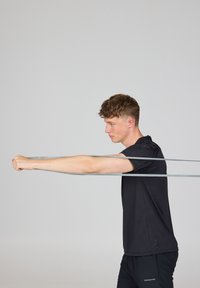 Jonge man in zwarte sportkleding die een grijze weerstandsband met beide armen naar voren strekt tegen een eenvoudige lichte achtergrond.