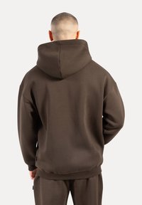 Muž s holou hlavou, oblečený v hnědé mikině a odpovídajících teplákách, zobrazený zezadu na jednobarevném pozadí.