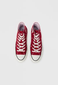 Κόκκινα καμβά παπούτσια ψηλής κορυφής με λευκές πουά τελείες, λευκά κορδόνια και μαύρες λαστιχένιες σόλες. Διαθέτουν λογότυπο σ το εξωτερικό μέρος.