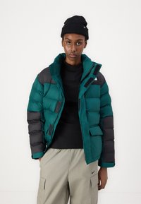 Pufová bunda v tmavom teal a čiernej farbe, so zvýšeným golierom, zapínaním na gombíky a zips, s textúrovaným povrchom a náprsníkovými vreckami.