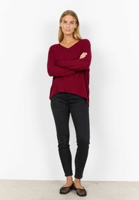 Bordeaux oversized v-hals trui met lange mouwen, gecombineerd met zwarte skinny jeans en zwarte mesh instapschoenen, gepresenteerd tegen een neutrale achtergrond.