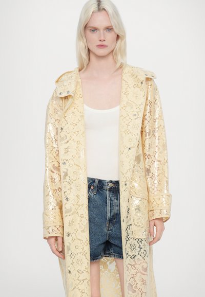 Manteau en dentelle jaune clair avec des sequins, doté d'un devant à boutons, d'un grand col et de poches latérales, porté sur un top blanc et un short en denim.