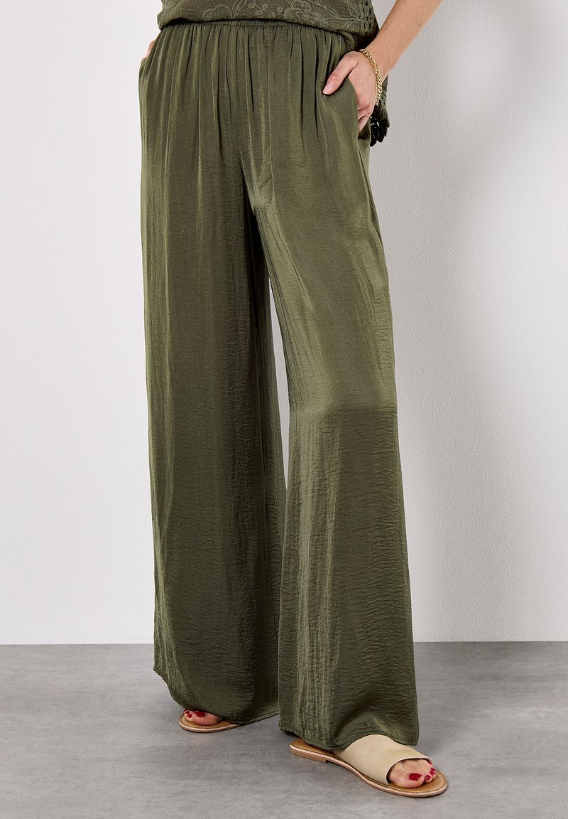 Une personne porte un pantalon large vert olive et un haut à motifs, se tenant sur un sol gris avec un mur de couleur claire en arrière-plan.