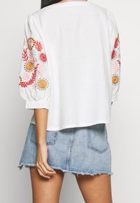 Blusa bianca con ricami floreali colorati sulle maniche, caratterizzata da maniche a sbuffo e una vestibilità comoda, abbinata a una gonna mini in denim.
