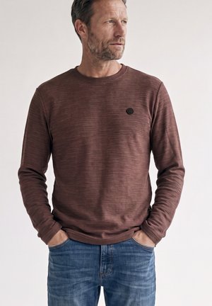 Middelaldrende mand med skæg iført en brun langærmet skjorte og blå jeans, hænderne i lommerne, ser til siden mod en ensfarvet baggrund.