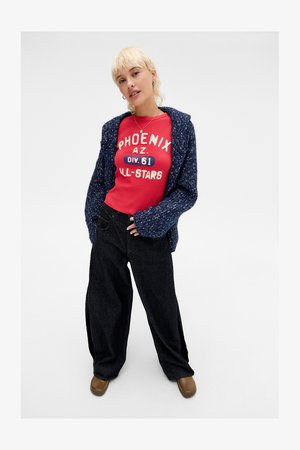 Jovem mulher com cabelo loiro, vestindo um cardigã azul de malha, uma camisa vermelha "Phoenix AZ", calças largas pretas e sapatos castanhos, a posar contra um fundo branco.