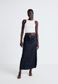 Jupe maxi en denim avec une coupe droite, en tissu bleu foncé, fermeture à boutons sur le devant, détails de couture visibles, portée avec un crop top blanc et des ballerines marron.