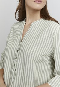 Blusa de lino de rayas verde claro y crema con escote en V, mangas cortas arremangadas y cuatro botones en la parte delantera. Textura suave y corte relajado.