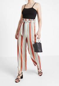 Pantalon large à rayures orange, blanc, bleu et vert avec un détail noué à la taille, assorti d'un haut noir sans manches et de sandales à fines lanières.