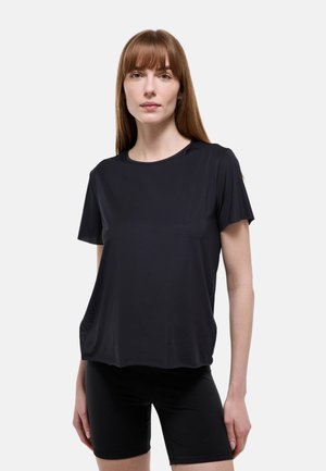Femme aux cheveux roux raides portant un t-shirt noir uni à manches courtes et un short de cyclisme noir, debout devant un fond clair.