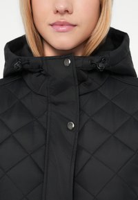Czarna pikowana kurtka z zapięciem na zamek, zatrzaskami oraz regulowanym kapturem. Gładki, lekki materiał z haftem w romby.
