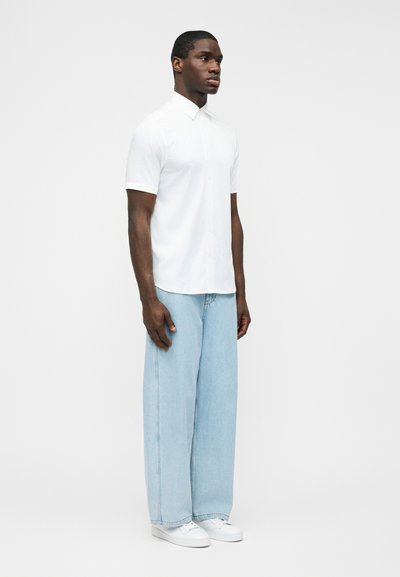 Άνδρας όρθιος, φοράει λευκό πουκάμισο με κοντά μανίκια και γιακά, ανοιχτό μπλε τζιν με φαρδιά παντελόνια και λευκά αθλητικά παπούτσια, με απλό φόντο.