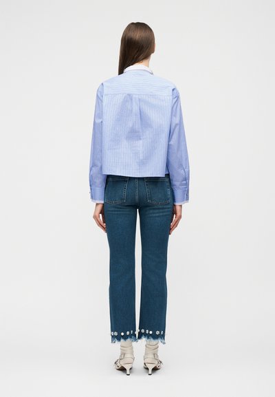 Γυναίκα που στέκεται πλάτη, φοράει μπλε ριγέ μπλούζα μακρυμάνικη, κοντά τζιν παντελόνια με ανθισμένες διακοσμήσεις και άσπρα σανδάλια με τακούνι.