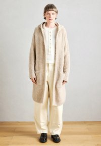 Veste longue beige texturée avec une capuche, dotée de deux poches avant. Portée sur un haut blanc et un pantalon crème clair, accompagnée de chaussures noires.