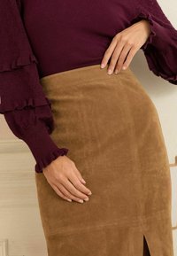 Burgundijas adīta blūze ar viļņotām piedurknēm, apvienota ar brūnu zamša zīda svārku, kuram ir sānu šķelšanās un gluda tekstūra.