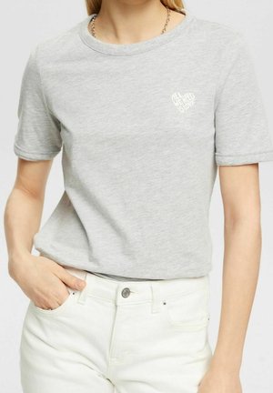 Vrouw draagt een lichtgrijs T-shirt met korte mouwen, met een klein wit hartje en de tekst "ALL WE NEED IS LOVE" op de borst.