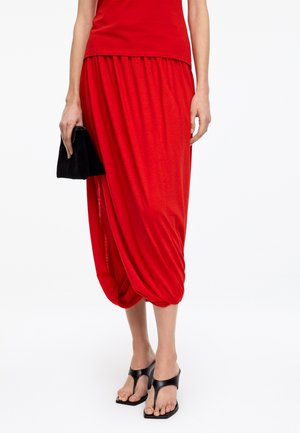 Femme portant une jupe midi drapée rouge, un haut rouge, des sandales à talons noires, tenant une petite pochette noire dans la main gauche.