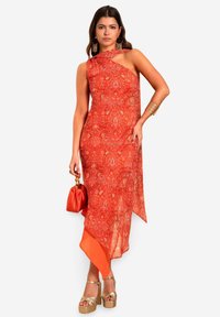 Vestido naranja asimétrico con tejido estampado, un hombro, y una textura suave y fluida, combinado con un bolso naranja y tacones de plataforma dorados.