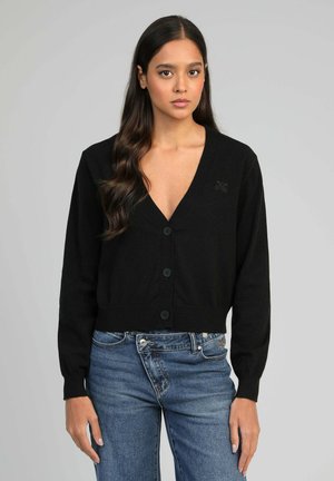Jeune femme aux longs cheveux bruns portant un cardigan noir à col en V boutonné et un jean bleu, se tenant devant un fond gris.
