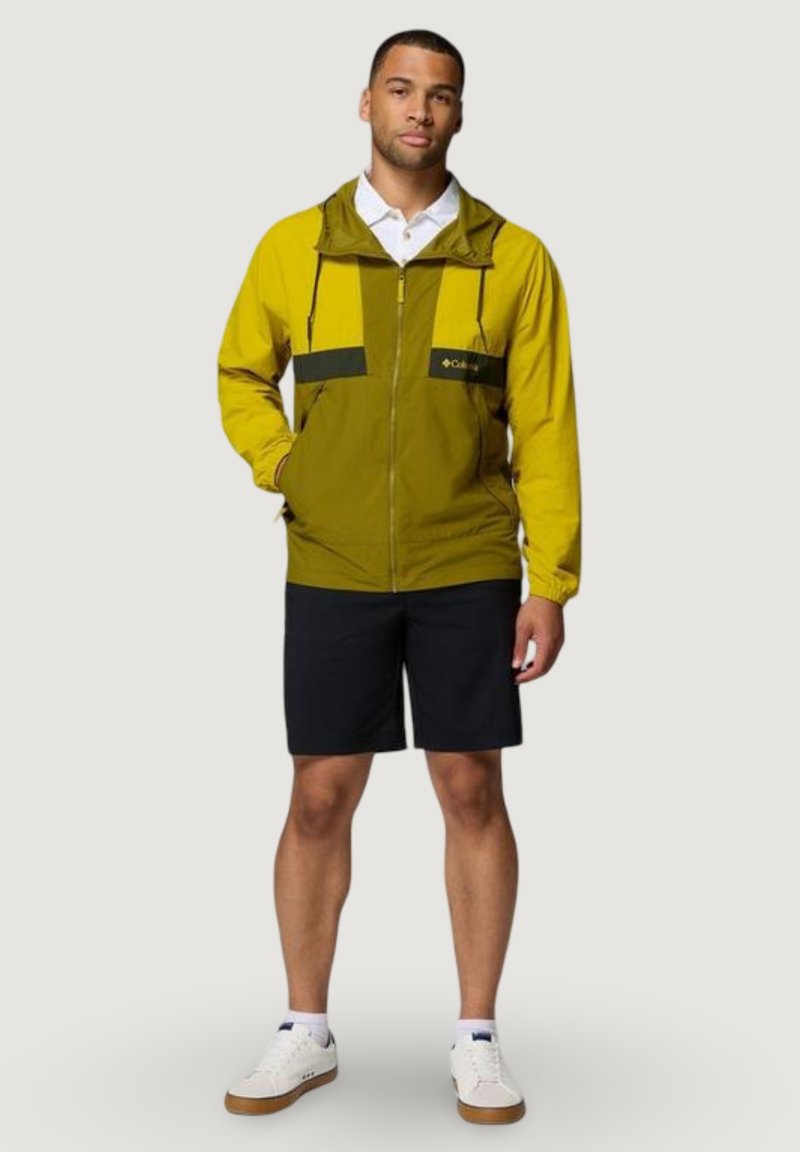 Hombre con chaqueta con cremallera amarilla y oliva, pantalones cortos negros, zapatillas blancas y camisa polo blanca sobre un fondo claro y liso.