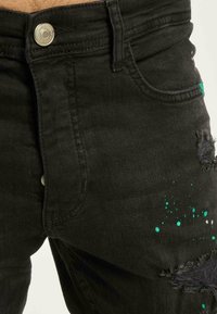 Črne denim hlače z raztrganimi detajli, z zapiranjem na sprednji gumb in teal barvnimi škropljaji na desni stegnu.