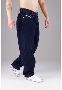 Dunkelblaue Denim-Jeans mit lockerem Schnitt, ausgestattet mit Kupfernieten, kontrastierenden Nähten und einem Lederpatch am hinteren Bund. Weiße Sneakers.