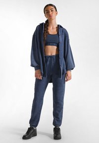 Marinblå loungewear-set bestående av en oversized skjorta med knappar, en kortad sportbh och byxor med snörning, tillsammans med svarta sneakers.