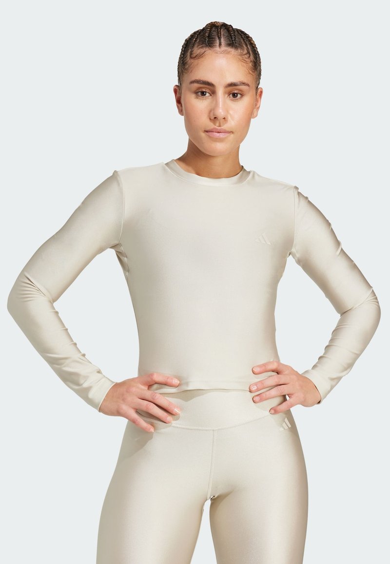 Långärmad ljusbeige träningsoverdel med en slät textur, rund halsringning och subtil logotypdetalj på bröstet, i kombination med matchande leggings.