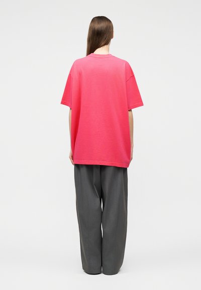 Άτομο με μακριά καστανά μαλλιά, ορατό από πίσω, που φοράει μια ροζ υπερμεγέθη κοντομάνικη μπλούζα και φαρδιά σκούρα γκρι παντελόνια, μπροστά σε απλό φόντο.