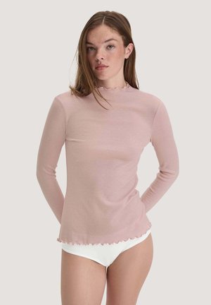 Junge Frau mit heller Haut und langen braunen Haaren, die ein blassrosa langärmliges Top und weiße Unterwäsche vor einem einfarbigen Hintergrund trägt.