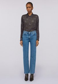 Frau in einem transparenten, dunklen Hemd mit Tiermotiven, hochgeschnittenen blauen Jeans und schwarzen Stiefeletten, die vor einem schlichten Hintergrund steht.