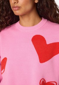 Ροζ πουλόβερ με μεγάλα κόκκινα σχέδια καρδιάς, κατασκευασμένο από απαλό πλεκτό υλικό με στρογγυλή λαιμόκοψη και υφασμάτινη υφή.