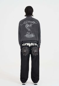 Osoba stoi tyłem do kamery, ubrana w szary sweter z wężem i napisem "Ed Hardy" oraz czarne spodnie z wzorem pajęczyny na tylnej kieszeni.