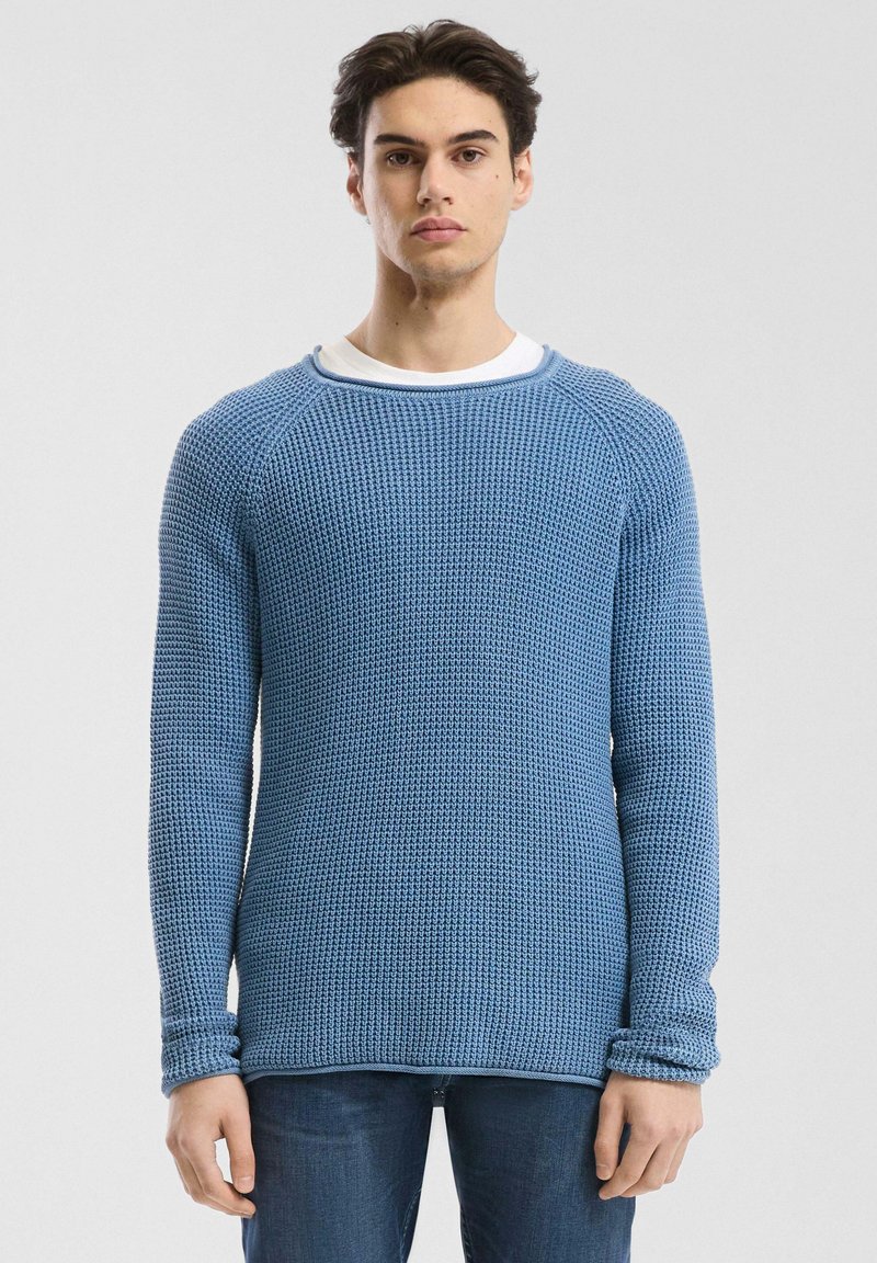 Helles blå, strukturiertes Strickoberteil mit langen Ärmeln, Rundhalsausschnitt und geripptem Saum, über einem weißen Hemd getragen, kombiniert mit dunkelblauen Jeans.