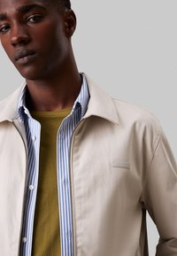 Giacca beige chiaro con zip, dal texture morbido, indossata sopra una maglietta verde e una camicia a righe blu. Presenta una toppa con logo sulla giacca.