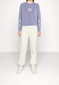 Femme portant un sweat-shirt lavande avec des motifs de bulles et le logo blanc Levi's, assorti à un pantalon de survêtement blanc et des baskets blanches.