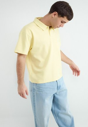 Junger Mann in einem hellgelben kurzärmeligen Poloshirt und hellblauen Jeans, der nach unten schaut, die Arme entspannt an den Seiten.