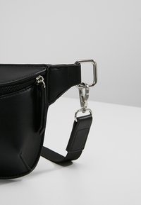 Sac banane en cuir noir avec une poche zippée, des ferrures argentées et une sangle amovible ornée d'un accent texturé noir.