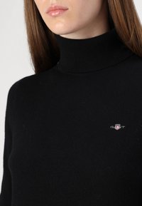 Pulover negru cu guler înalt, realizat dintr-un material tricotat moale. Prezintă un logo brodat mic pe piept și un design ajustat în jurul gâtului.