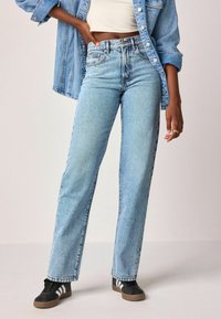Jeans de denim azul claro con corte recto, cintura alta y textura desgastada, combinados con zapatillas deportivas en blanco y negro.