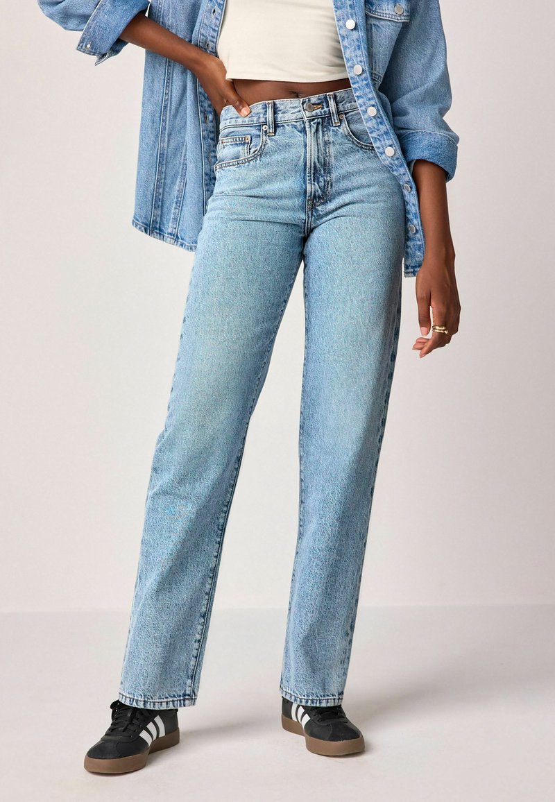 Jeans de denim azul claro con corte recto, cintura alta y textura desgastada, combinados con zapatillas deportivas en blanco y negro.