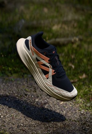 Musta-beige Salomon juoksukenkä, jossa on oransseja yksityiskohtia, leijuu soratiellä ulkona.
