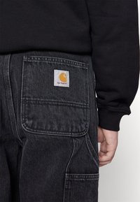 Los tirantes de mezclilla oscura presentan un gran bolsillo trasero con una etiqueta blanca que muestra un logo amarillo. La manga de una camisa negra es visible.