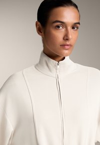 Lys beige zip-up sweatshirt med høj krave, blød tekstur og lodrette sømdetaljer foran. Rent og minimalistisk design.