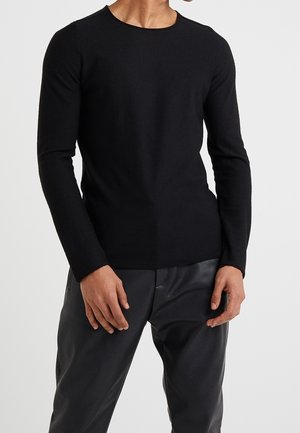 Mann trägt ein eng anliegendes, einfarbiges schwarzes Langarmshirt und schwarze Hose, steht mit entspannten Händen seitlich an einem weißen Hintergrund.